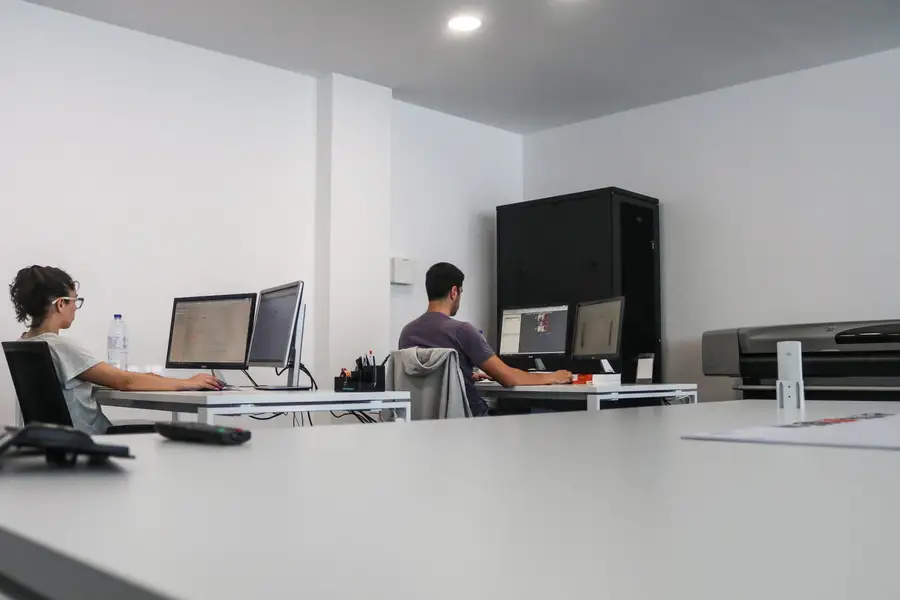Dois projetistas em frente aos monitores, mesa de trabalho branca em primeiro plano